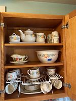 Wooden cabinet shelf with Corelle Abundance patterned stoneware including teapot, mugs, cream and sugar set, large bowl, and saucers.