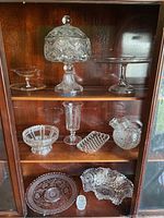 Wide view showing various cut and pressed glass serving pieces including the lidded pedestal bowl, pitcher, pedestal plate, and additional glassware items in a wooden cabinet.