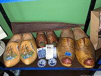 Photo of three pairs of wooden shoes with different painted designs or plain, two small Delft blue and white ceramics, two blue and white tiles, and a wooden boat tray in the background.