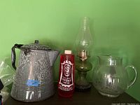 Photo showing enamelware kettle, red lamp oil bottle, green glass oil lamp, and etched glass pitcher against a green wall background.