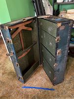 Open view of Girard College steamer trunk showing four wooden drawers with ring pulls on the right side, wooden hangers and hanging supports on the left, interior patterned lining, and metal and leather exterior edge reinforcements.
