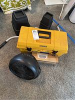View of yellow Hechbrenner toolbox, black portable fan, black plastic furniture risers+bed risers, and white PVC elbow pipe on carpeted floor.