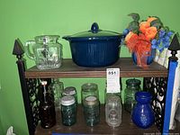 Photo showing multiple glass jars, blue and clear, some with zinc and glass lids, a dark blue lidded crock, a clear glass pitcher with handle and geometric design, a blue glass vase, and a tall brown bottle on a wooden shelf against green wall.