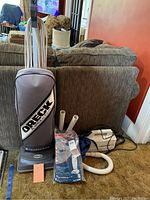 Full view of lot showing Oreck XL Shield upright vacuum, handheld vacuum, packaged bags, accessory hoses, all beside a couch.