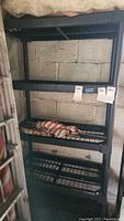 Photo of black plastic shelving unit with four grid shelves against a concrete block wall, showing empty shelves and size tags.