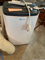 Front view of Rollibot portable air conditioner showing white and gray body, exhaust hose attached, power cord draped over top and remote control placed on lid