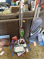 Shark Sonic Duo floor scrubber standing upright with cleaning pads, bottles of cleaning solution, and other cleaning tools around it.
