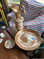 Set of three items on wooden table near window: two decorated Asian style plates and one tall ornate vintage pitcher with bird-shaped handle.