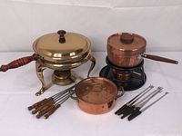 Overview of lot showing large covered chafing dish with wooden handle, copper fondue pot on burner stand with wooden handle, small covered copper pot, and fondue forks and skewers with wooden handles.