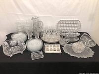 Wide overview of entire glass and crystal lot on black cloth, showing the variety and arrangement of items including pitcher, glasses, plates, and trays.