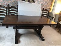 Front angle view of vintage wooden trestle dining table on carpet with floral wallpaper background, showing the polished surface and pedestal base.