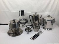 Full view of the entertaining lot items on white draped background: covered chafing dish, Pyrex coffee pot with warming stand, kettle, ice bucket, candle stick holder, metal bowl, and cocktail forks.