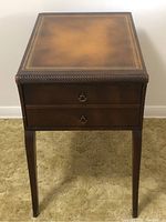 Front view of vintage end table showing leather top surface, two drawers with brass handles, and tapered legs on carpeted floor.