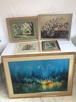 Five framed artworks displayed on a white table and against a white wall, showing different sizes and styles including abstract, landscape, and floral themes.