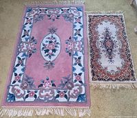 Two wool rugs side by side on floor: larger pink with floral border and central medallion, smaller beige with intricate central and border patterns, both with fringed edges.