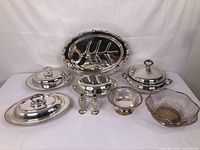 Full lot view showing two covered oval serving dishes with knob handles on lids, pair of salt and pepper shakers, oval serving tray with decorated edge, silver plated pierced basket bowl, wire framed bowl with tarnished metal base, and small covered dish.