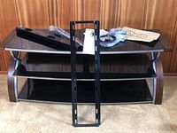 Full view of the Whalen 3-in-1 TV stand showing three black tempered glass shelves supported by curved dark wood or wood-like side panels. Mounting bracket and accessory cables rest on top shelf.