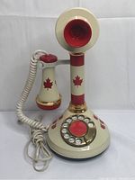 Front view of the vintage DecoTel candlestick telephone decorated with Canadian maple leaf icons, showing the handset resting on its cradle and the rotary dial in front.