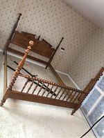 Wooden four-post bed frame set up leaning in a room with vintage wallpaper, showing headboard, footboard with turned posts and spindles, and metal support rails on the floor behind the frame.