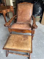 Front view of antique wooden birthing chair with attached arms and original leather seat. Ottoman in front with matching leather top and nail-head trim.