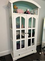 Full view of white armoire cabinet with glass windows and bottom drawer, displaying folded clothing and items inside.
