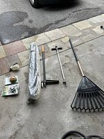 Photo showing assorted garden tools including two hand weeders, a black plastic leaf rake, a wrapped long item in plastic, a handheld sprayer, and some small metal garden tools.