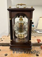 Front view of wooden mantel clock with glass panels, showing clock face with Roman numerals and pendulum.