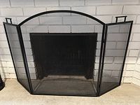 Front view of the black metal mesh folding fireplace screen with three panels expanded in front of a brick fireplace.