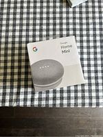 Boxed Google Home Mini on a checkered fabric surface, showing front view of packaging.
