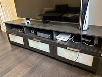 Full view of IKEA TV console with a large flat-screen TV on top, showing three drawers and open compartments with gaming consoles and accessories.