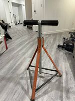 Full view of the adjustable out feed roller showing the orange triangular metal base, vertical support pipe with measurement markings, and black rubber roller on top.