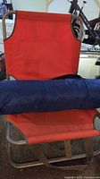 Red folding beach chair with metal frame and blue carrying bag for sun shade tent placed horizontally on the chair seat.