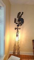 Full view of tall metal floor lamp with rooster weather vane on top, lit and placed against wall corner.