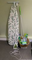 Full sized ironing board with leaf patterned cover leaning against wall, green and white T-Fal steam iron on floor, green-handled Swiffer mop, woven basket with cleaning supplies