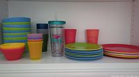 Full view of colorful plastic plates, bowls, cups and a clear plastic tumbler with straw inside a white shelf