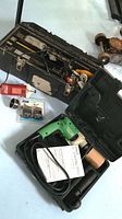 Photo of Hammer Drill in black-green hard plastic case with instruction manual visible, with open toolbox near it containing hand tools