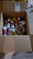 Box of mixed used golf balls showing white, yellow, orange, and pink colored golf balls with visible brand logos and various dimple patterns.