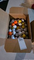 Opened cardboard box filled with a variety of golf balls showing multiple colors including orange, yellow, white.