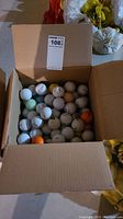 Overview photo of cardboard box filled with various golf balls, mostly white with a few colored ones like orange and green.