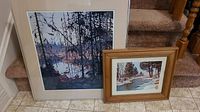 Two framed and matted prints on stairs, a larger 24x22 inch print depicting a forest and lake scene and a smaller 14x16 inch print depicting a snowy creek scene.