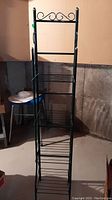 Front view of a tall black metal shelf stand with four wire shelves and decorative curved metal accents at the top
