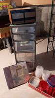 Tall plastic storage bin with five clear drawers on wheels, black storage box on top, scrapbook kit, and plastic storage containers on floor in basement setting.
