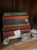 A collection of six antique books in varied condition stacked horizontally, a metal chess clock, and two metal decorative items on a wooden shelf.