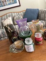 Photo showing assortment of glass vases in various colors including clear, purple, green, amber, and a decorative skull-shaped glass candle holder along with small candles in glass jars.