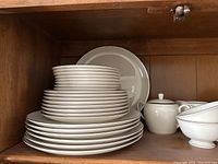 Shelved white china set including stacked plates of multiple sizes, sugar container, and part of the cup set visible