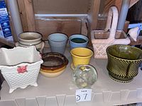 Various small flower pots in different colors, shapes, and materials arranged on shelf including ceramics, pottery, and glass decorative item.