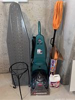 Photo showing the metal ironing board, Bissell vacuum cleaner, orange mop, small black metal table, and carpet detergent jug all together against a concrete wall.