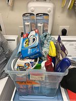 Plastic bin filled with various laundry and household cleaning items showing used detergent, disinfectant, utility lights, and other products.