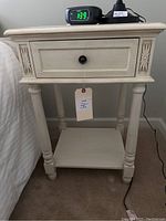 Front view of the wooden nightstand showing drawer with black knob, floral carvings, turned legs, lower shelf, and items on top.