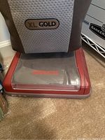 Close-up photo of the bottom part of the Oreck XL Gold upright vacuum showing the red base with an Oreck logo, and the grey and brown vacuum bag with XL Gold label.